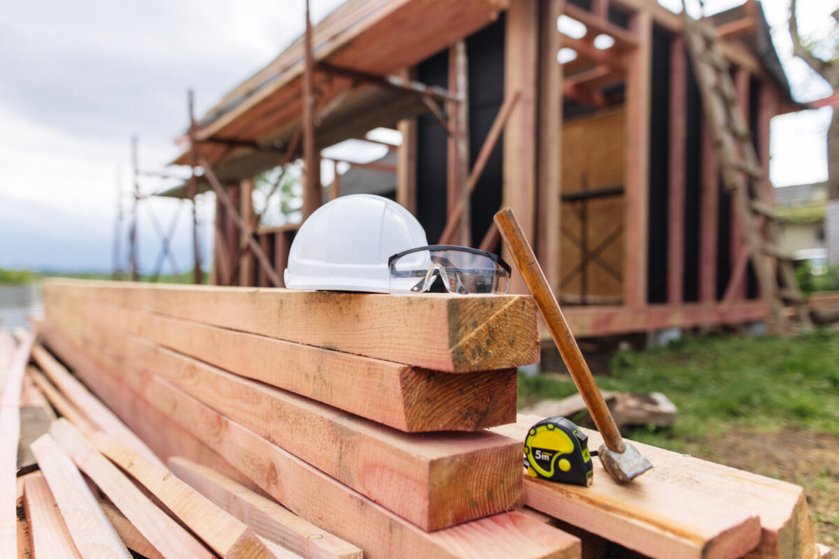 The protective helmet, glasses and hammer lie on the stacked lumber at the frame house construction site