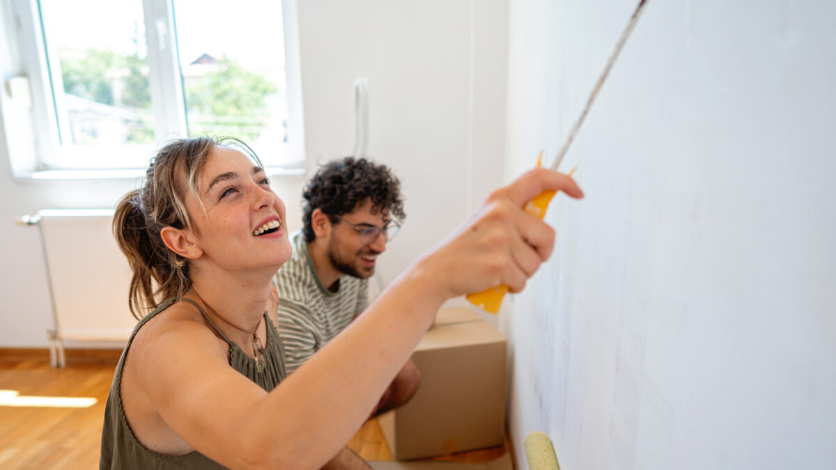 Kneeling side by side with rollers in hand, a couple shares a lighthearted moment while painting. The warm tones of the wooden floor and bright window create a cozy, inviting atmosphere in their apartment.