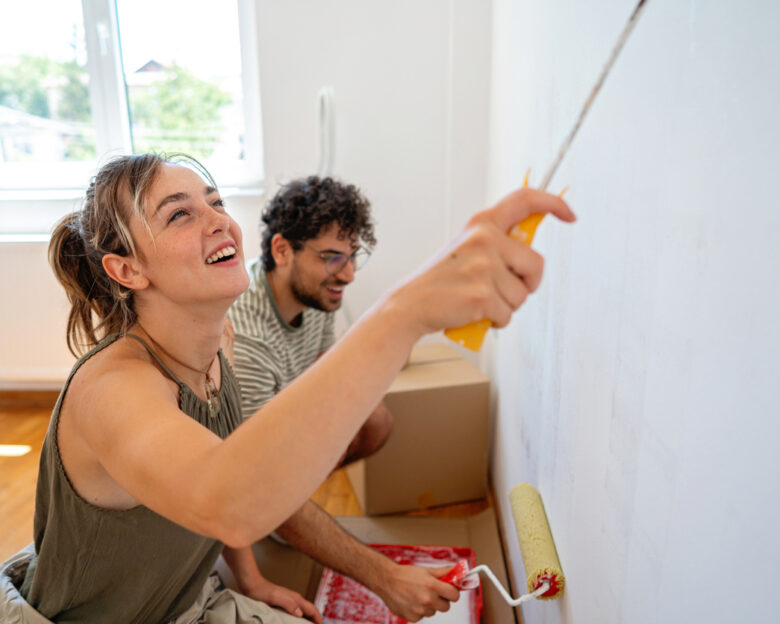 Kneeling side by side with rollers in hand, a couple shares a lighthearted moment while painting. The warm tones of the wooden floor and bright window create a cozy, inviting atmosphere in their apartment.
