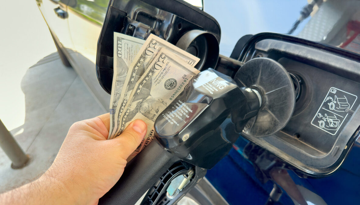 Long SEO Description A close-up conceptual photo shows a person's hand inserting a stack of US twenty dollar bills into a gas pump nozzle instead of a credit card, attached to a blue car at a fuel station under bright daylight. This symbolic image powerfully represents the soaring gasoline prices, increasing fuel expenses, and economic strain on drivers due to high energy costs in the United States, with full frame composition and copy space available for text or graphics. Ideal for editorial coverage on inflation, oil prices, consumer spending, transportation budgets, energy crisis topics, or financial impact illustrations in news articles, blog posts, social media campaigns, and business reports.