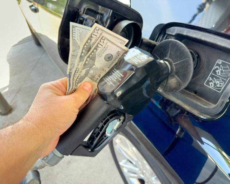 Long SEO Description A close-up conceptual photo shows a person's hand inserting a stack of US twenty dollar bills into a gas pump nozzle instead of a credit card, attached to a blue car at a fuel station under bright daylight. This symbolic image powerfully represents the soaring gasoline prices, increasing fuel expenses, and economic strain on drivers due to high energy costs in the United States, with full frame composition and copy space available for text or graphics. Ideal for editorial coverage on inflation, oil prices, consumer spending, transportation budgets, energy crisis topics, or financial impact illustrations in news articles, blog posts, social media campaigns, and business reports.