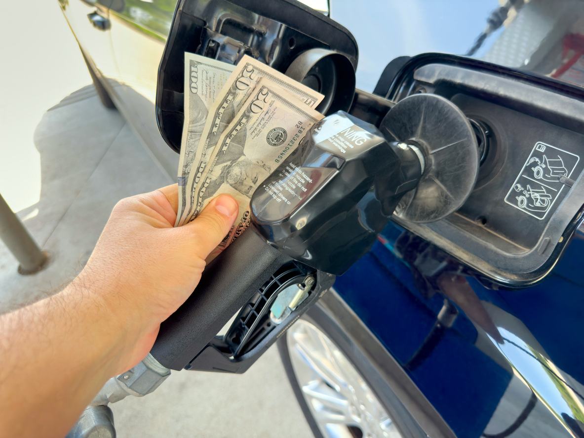 Long SEO Description A close-up conceptual photo shows a person's hand inserting a stack of US twenty dollar bills into a gas pump nozzle instead of a credit card, attached to a blue car at a fuel station under bright daylight. This symbolic image powerfully represents the soaring gasoline prices, increasing fuel expenses, and economic strain on drivers due to high energy costs in the United States, with full frame composition and copy space available for text or graphics. Ideal for editorial coverage on inflation, oil prices, consumer spending, transportation budgets, energy crisis topics, or financial impact illustrations in news articles, blog posts, social media campaigns, and business reports.