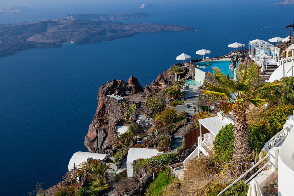 Scenic high up view from Firostefani, Santorini, with terrace pool, palm tree, volcanic cliffs, and calm blue sea.