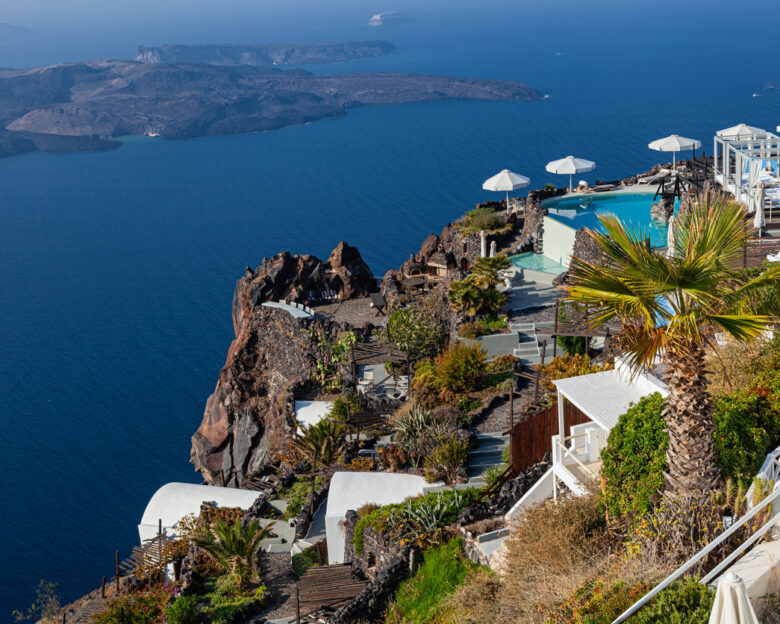 Scenic high up view from Firostefani, Santorini, with terrace pool, palm tree, volcanic cliffs, and calm blue sea.