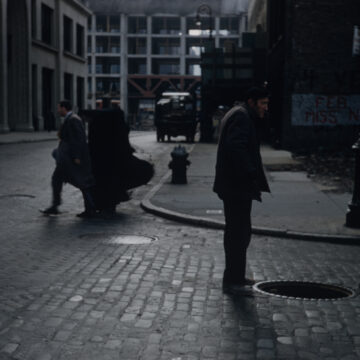 A man in a cap stand at the edge of an open manhole on a brick-lined street in Tribeca, New York