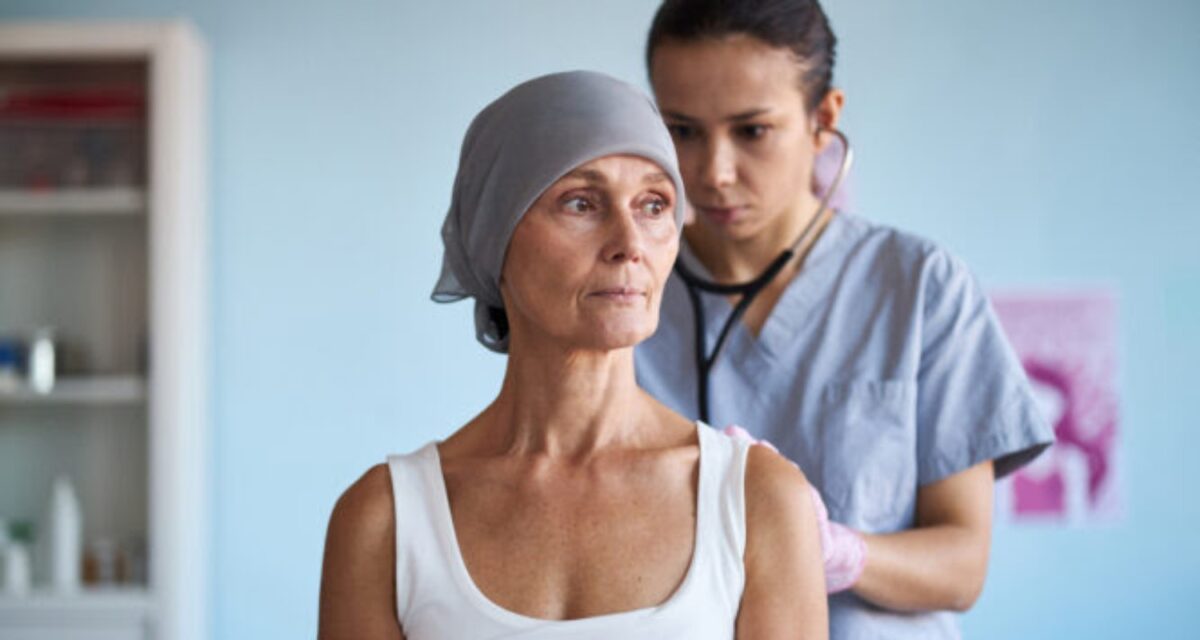 Middle aged Caucasian woman wearing headscarf sitting while young adult female doctor examining patient with stethoscope in medical setting related to breast cancer treatment