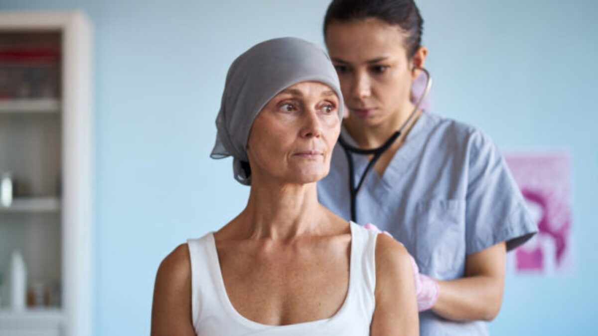 Middle aged Caucasian woman wearing headscarf sitting while young adult female doctor examining patient with stethoscope in medical setting related to breast cancer treatment