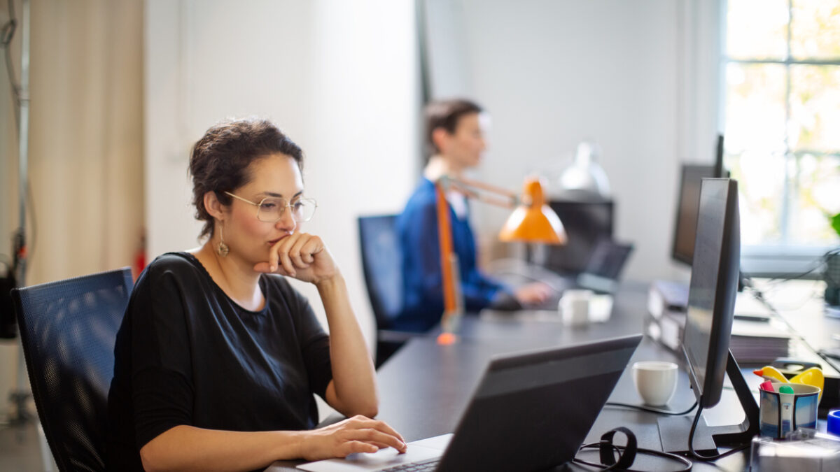 Businesswoman working on laptop with colleague in background. Female executive sitting at her desk looking busy while working on laptop computer.