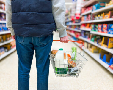 Low angle close up color image depicting a man holding a shopping basked filled with essential fresh groceries like bread and milk in the supermarket.