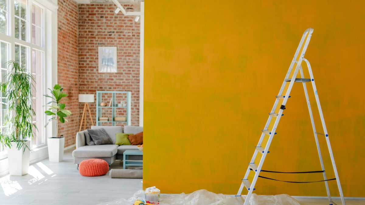 Ladder placed beside a vibrant yellow wall mid-paint, with a comfy living space and decorative shelving visible behind.