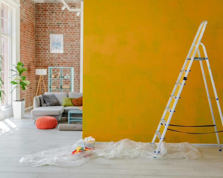 Ladder placed beside a vibrant yellow wall mid-paint, with a comfy living space and decorative shelving visible behind.
