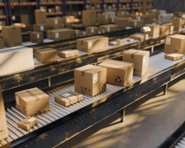 Rows of cardboard boxes and parcels travel along conveyor belts inside a large distribution warehouse, illustrating efficient shipping, order fulfillment, inventory handling and modern logistics operations.