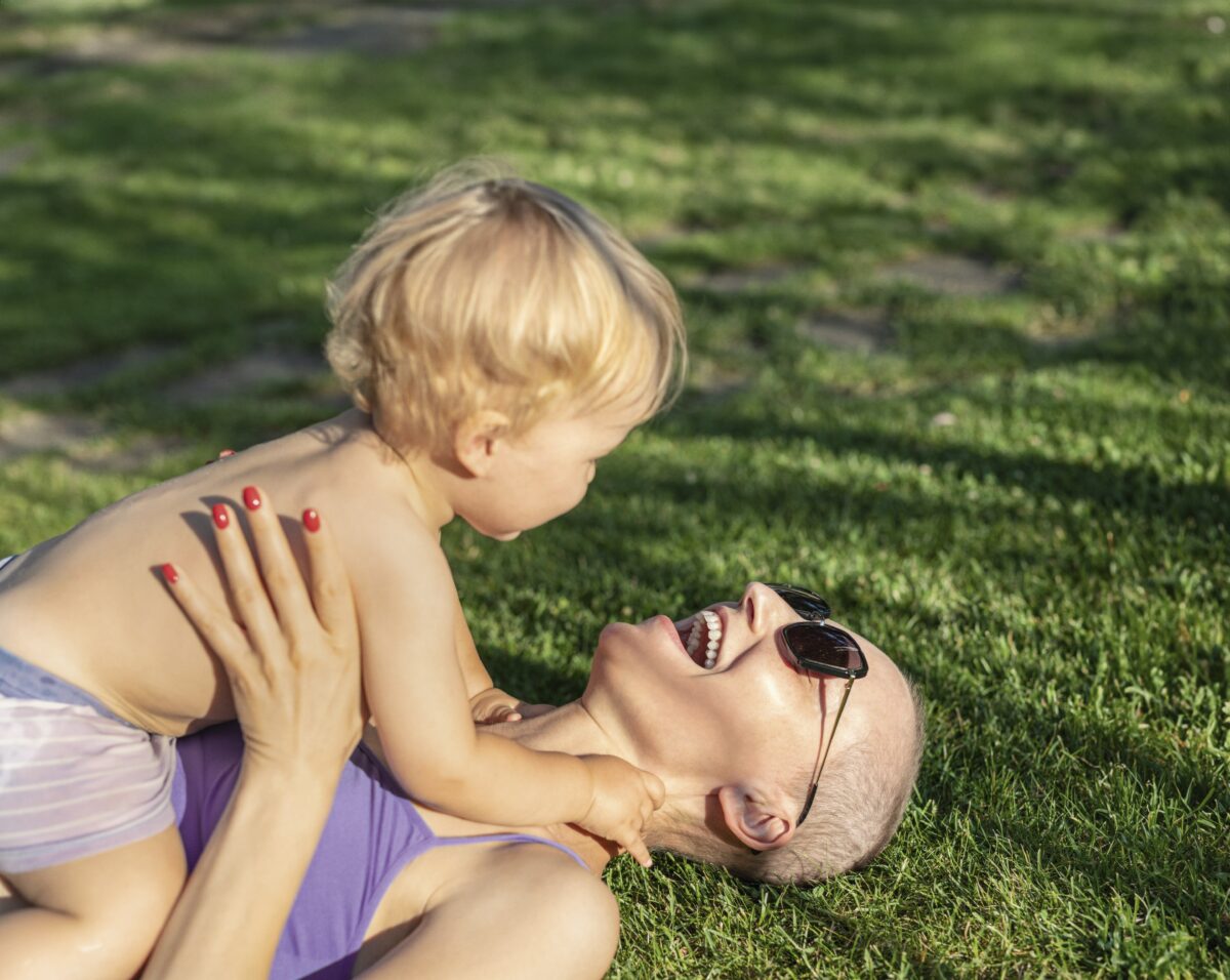 Young adult beautiful bald caucasian mom enjoy lying with little blond toddler boy son on green grass lawn at city park at summer day. Mother happy parenting and childcare . Healthy baby outdoors.