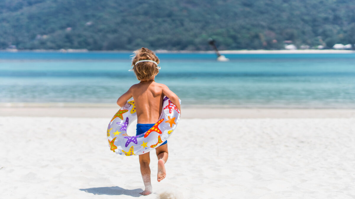 Back view of little boy with swimming tube running towards the sea in summer day. Copy space.