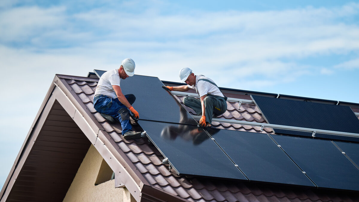 Workers building solar panel system on rooftop of house. Two men installers in helmets installing photovoltaic solar module outdoors. Alternative, green and renewable energy generation concept.