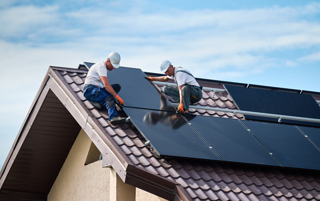 Workers building solar panel system on rooftop of house. Two men installers in helmets installing photovoltaic solar module outdoors. Alternative, green and renewable energy generation concept.