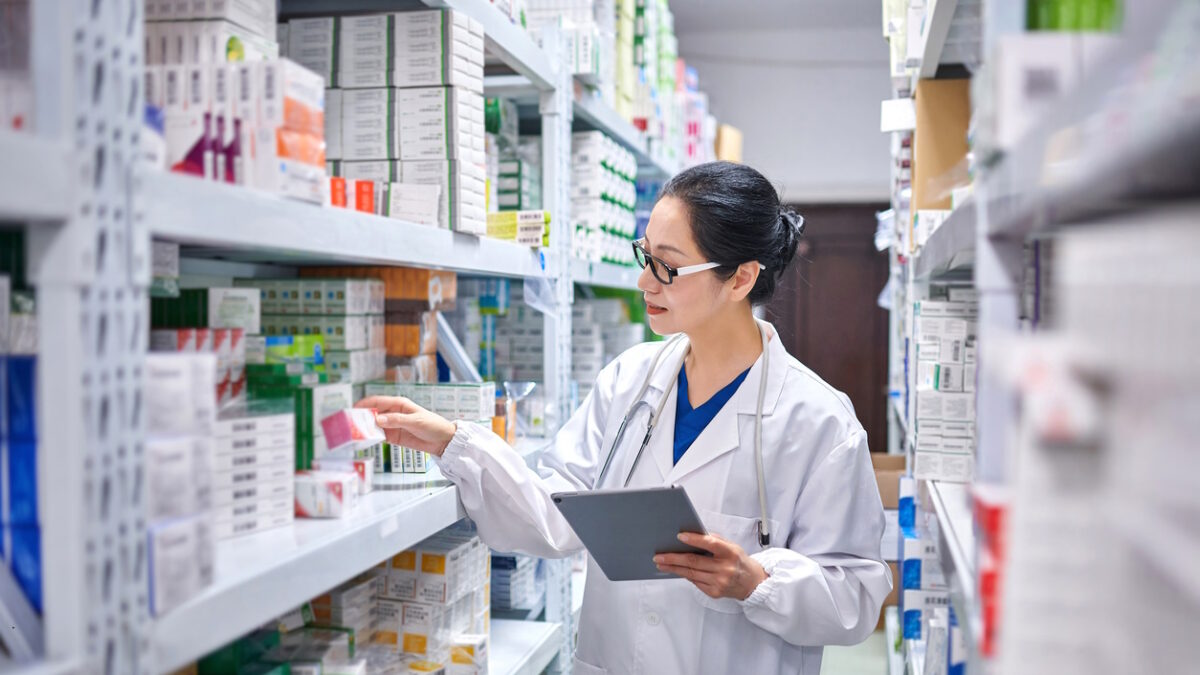 Side view of one asia female pharmacist using digital tablet while taking inventory at pharmacy warehouse.