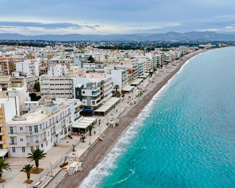 An aerial view of Loutraki, a charming coastal town in Greece known for its scenic waterfront promenade and vibrant urban landscape. The image highlights the turquoise waters of the Corinthian Gulf, lined with modern buildings, green parks, and palm trees, creating a perfect blend of natural beauty and urban charm. Loutraki is renowned for its thermal spas, cultural heritage, and inviting beaches, making it a popular destination for tourists seeking relaxation and leisure. The town's unique location provides breathtaking views of the sea and surrounding mountains, ideal for travel and lifestyle themes.