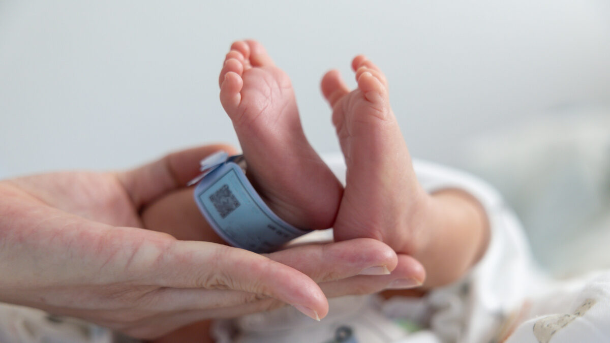A baby's feet are held up by a person. The baby is wearing a blue tag. The baby is in a hospital bed
