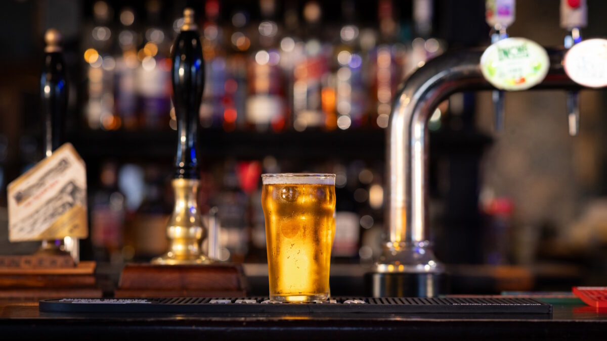 A full shot of a full pint of lager on a bar counter. The pint is overflowing with beer foam. The rustic pub is located in Hexham.