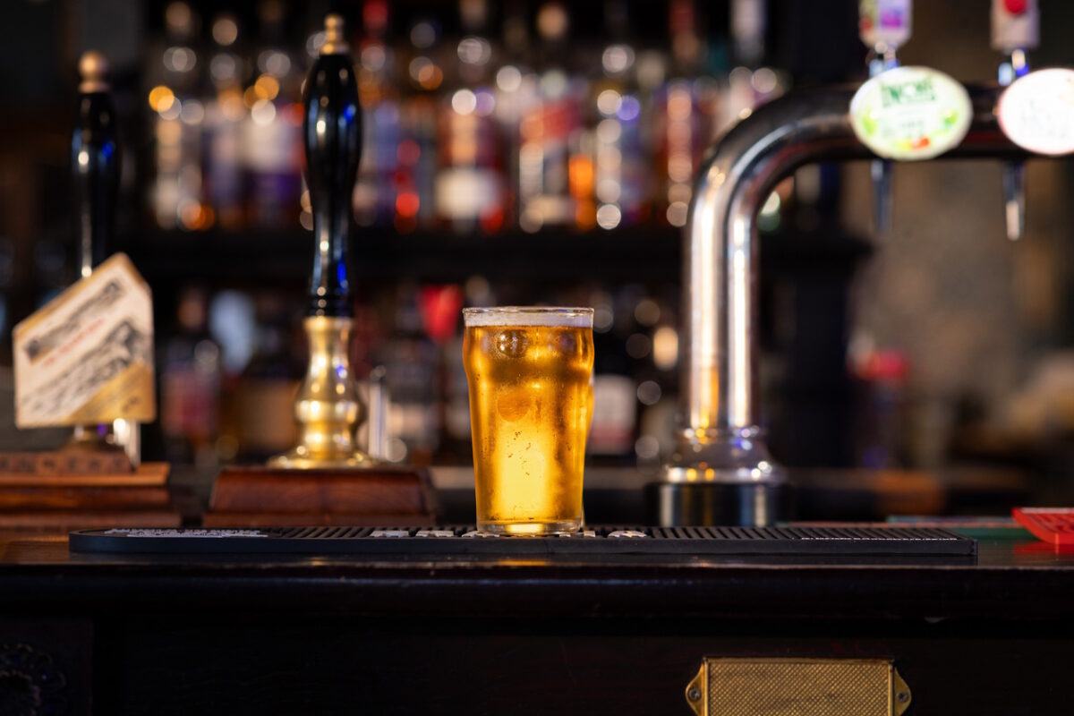 A full shot of a full pint of lager on a bar counter. The pint is overflowing with beer foam. The rustic pub is located in Hexham.