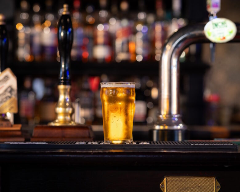 A full shot of a full pint of lager on a bar counter. The pint is overflowing with beer foam. The rustic pub is located in Hexham.