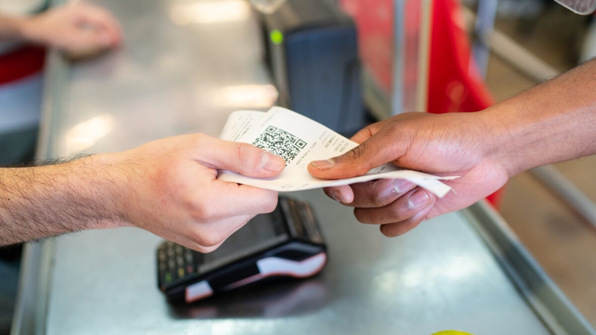 Hands exchange a receipt at a supermarket checkout counter after a grocery purchase. Two people finalize a transaction exchanging a paper slip containing a QR code at grocery store checkout counter.