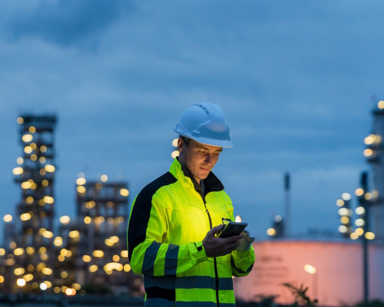 Industrial worker using a smartphone for communication at an oil and gas plant at night.