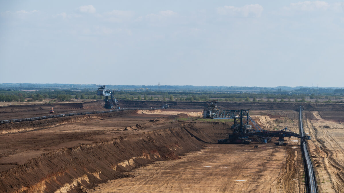 A vast open pit mining site featuring large blue excavators and extensive conveyor belt systems. The layered soil formations and industrial machinery highlight the scale of mineral extraction under a clear sky.
