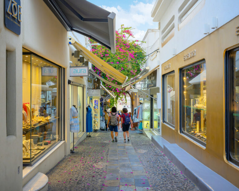 Pedestrians shop on the narrow cobblestone Ipapantis shopping street, nicknamed Gold Street, lined with boutiques, jewelry stores and bougainvillea plants in Fira, the capital town of Santorini island, Greece.