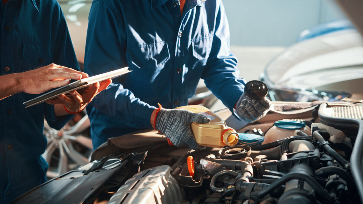 Mechanic pouring oil in car engine when his colleague using tablet computer