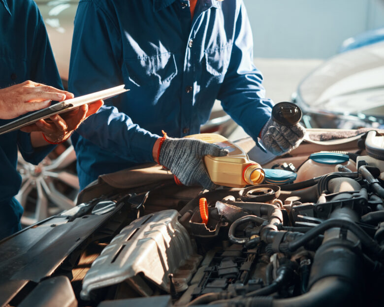 Mechanic pouring oil in car engine when his colleague using tablet computer