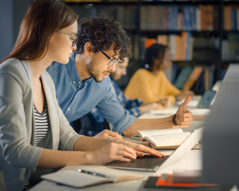 University Library: Talented Caucasian Girl uses Laptop, Smart Helpful Classmate Explains and Advices Her with Class Assignment. Happy Diverse Students Talking, Learning, Studying Together for Exams