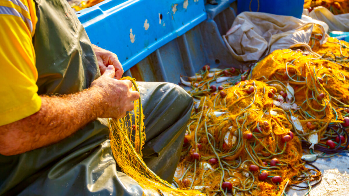 Fisher in rubber trousers and boot siting in his boat and pile up fishing net for angling at open sea.