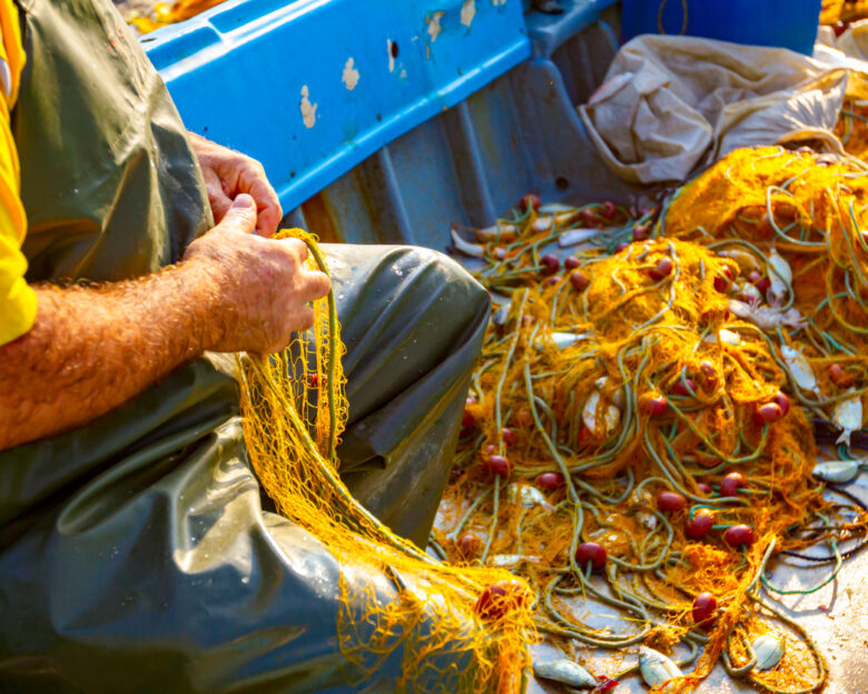 Fisher in rubber trousers and boot siting in his boat and pile up fishing net for angling at open sea.