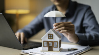 Close-up of a tiny house shielded by an umbrella atop loan documents next to keys and a laptop, representing property coverage.