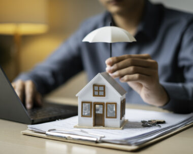 Close-up of a tiny house shielded by an umbrella atop loan documents next to keys and a laptop, representing property coverage.