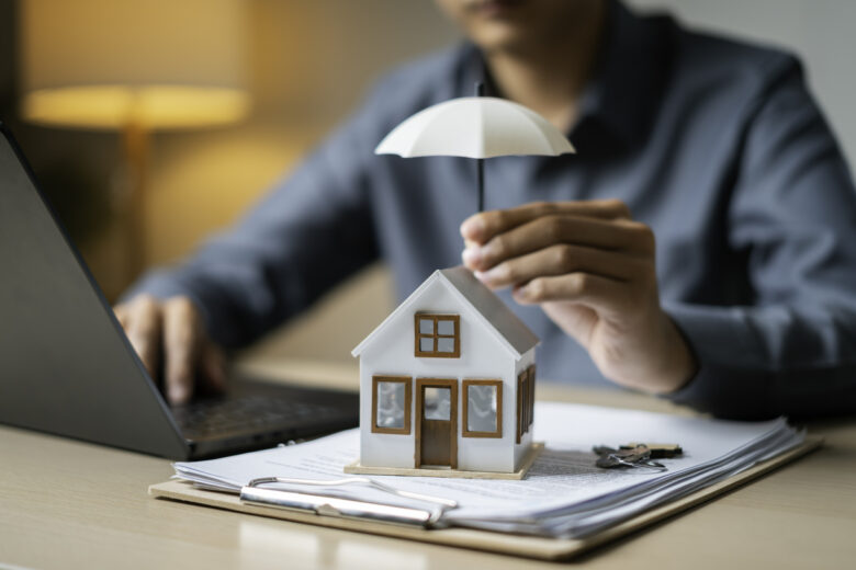 Close-up of a tiny house shielded by an umbrella atop loan documents next to keys and a laptop, representing property coverage.