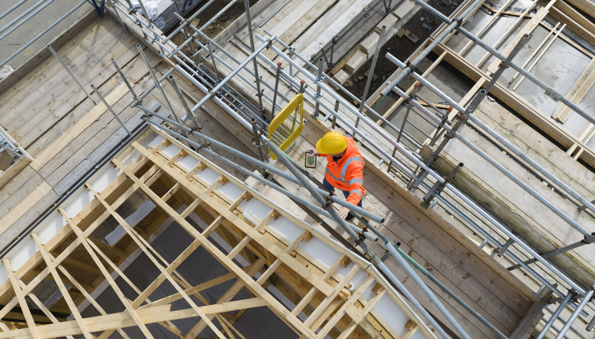 Construction worker on building site showing housebuilding, roof trusses, scaffolding and completed houses. Cornwall, UK