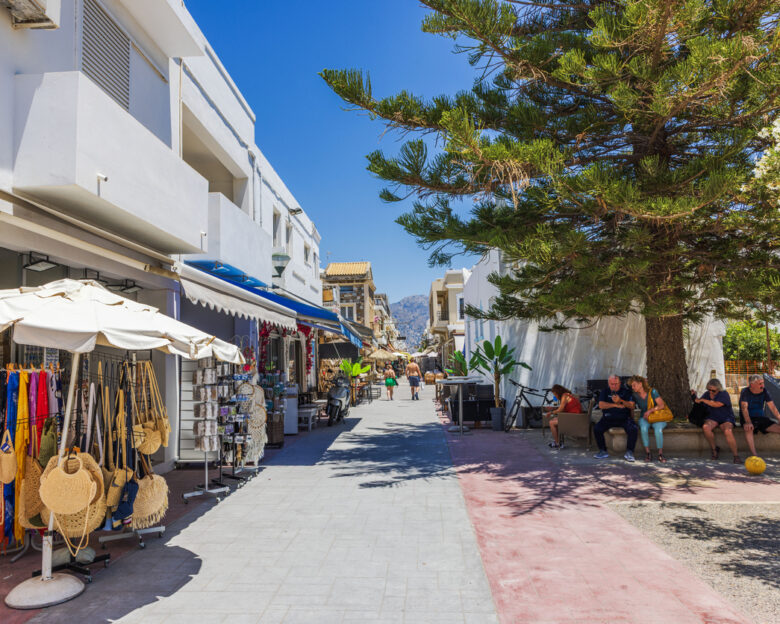 Kardamena. Greece. July.07.2025. Beautiful view of Mediterranean street with shops souvenirs and people relaxing in shade on background of mountains in sunny day.