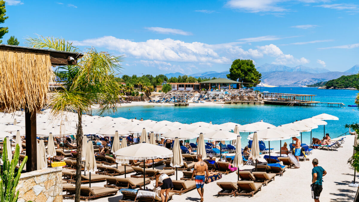 Ksamil, Rruga Riviera - Rows of brown sunbeds and white umbrellas fill a sandy beach club with a thatched-roof structure in the foreground. A turquoise bay and distant mountains are visible beyond.