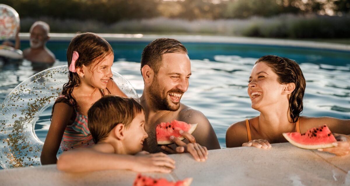 Happy parents and their kids talking while eating watermelon during summer day in the swimming pool.
