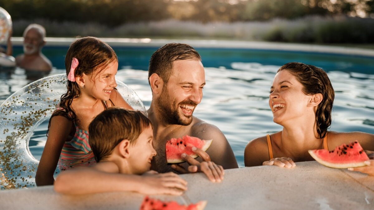 Happy parents and their kids talking while eating watermelon during summer day in the swimming pool.