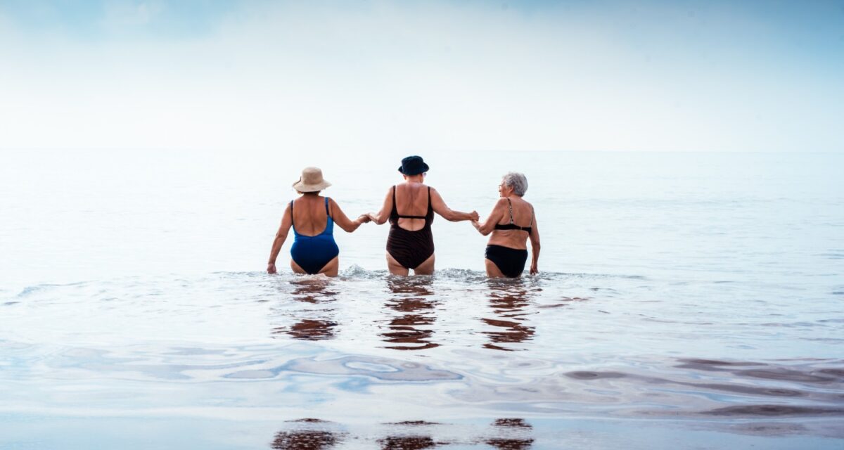 Three mature women friends holding hands as they wade into calm ocean waters on a sunny beach vacation, enjoying companionship, freedom and active retirement together