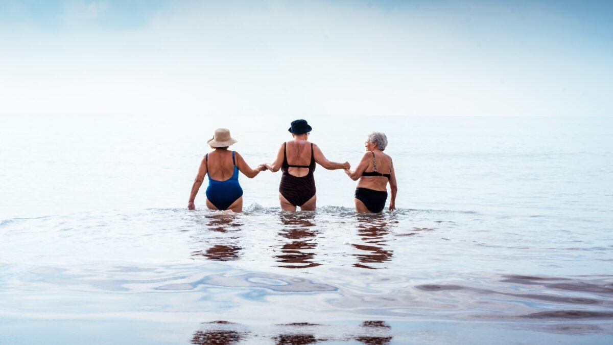 Three mature women friends holding hands as they wade into calm ocean waters on a sunny beach vacation, enjoying companionship, freedom and active retirement together