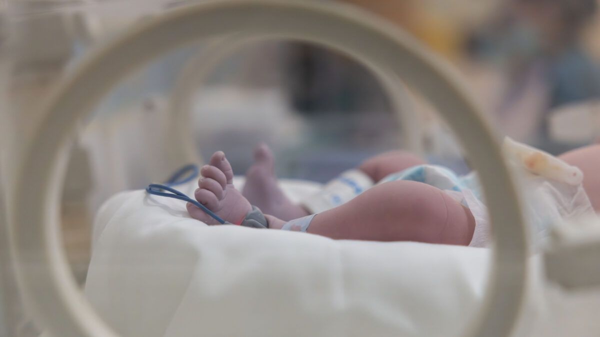 A baby's feet are visible in a hospital incubator. The baby is wrapped in a blanket and has a blue strap around its foot. The baby is in a hospital setting, and the image conveys a sense of care