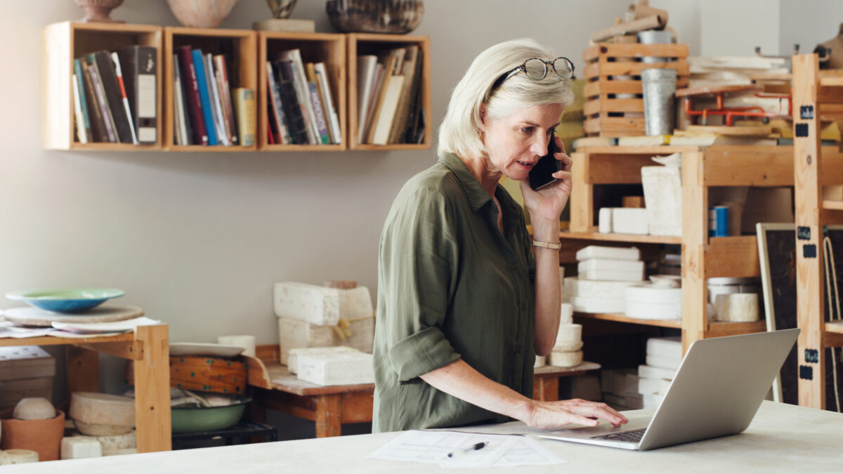 Shot of a mature woman talking on a cellphone and using a laptop while working in a pottery studio