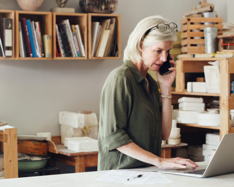 Shot of a mature woman talking on a cellphone and using a laptop while working in a pottery studio