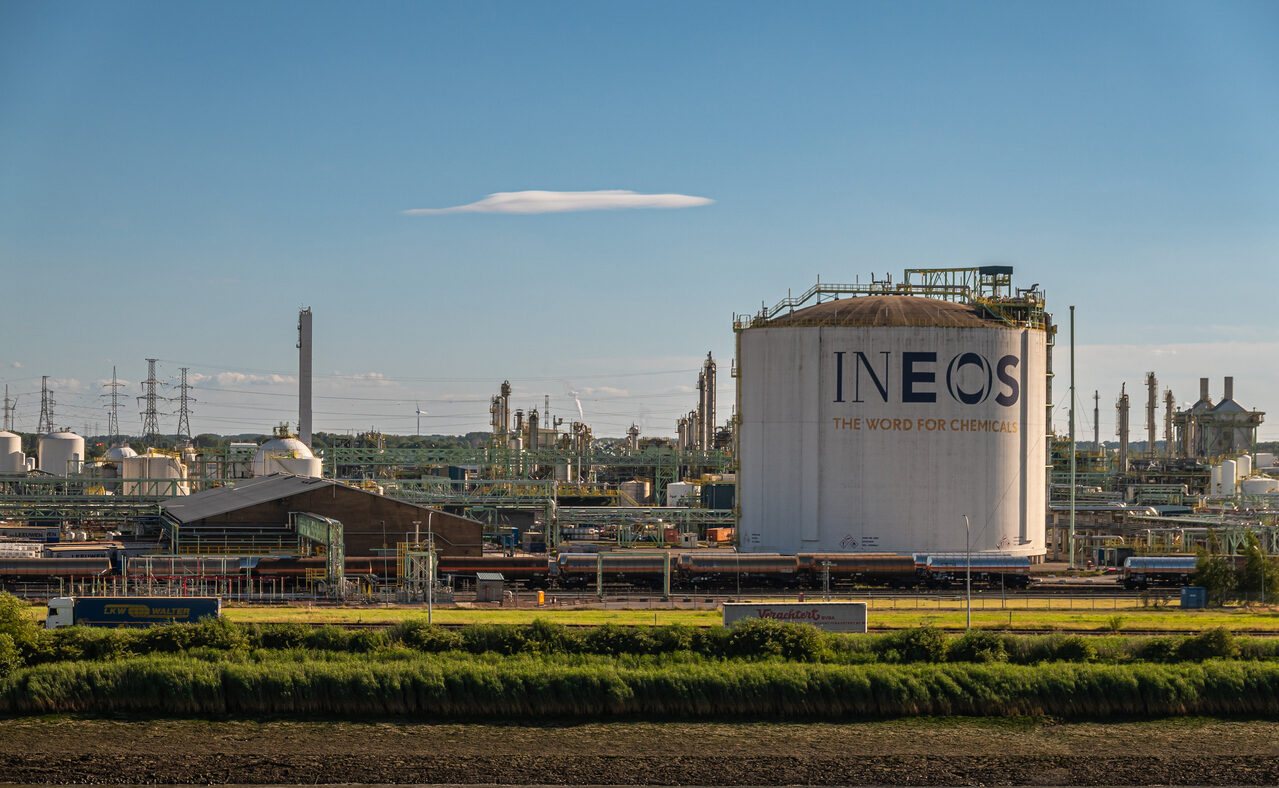 Antwerpen, Flanders, Belgium - July 10, 2022: Chemical Industry along left-shore Scheldt River. INEOS Zwijndrecht large storage tank with sign and slogan under blue sky. Green shoreline