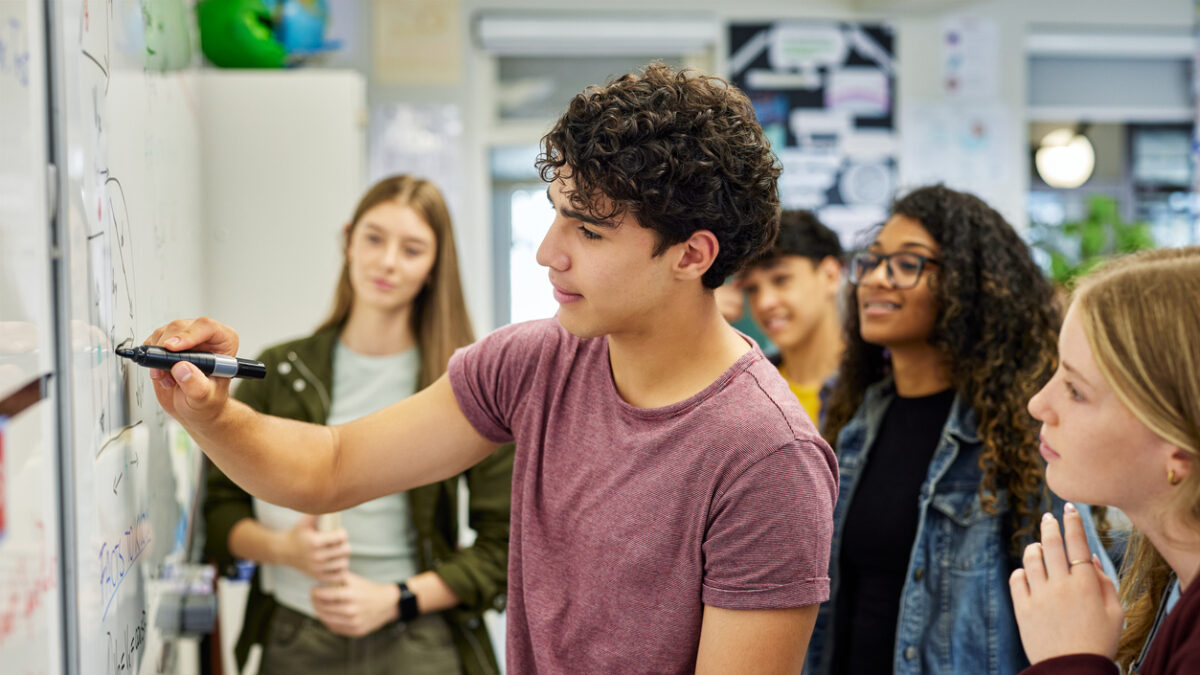 Male student writing on whiteboard while explaining a concept to his classmates at secondary school. Focused teens delivers a group presentation while drawing on board as classmates observe attentively. Teen guy helping classmates understand solution to geometry or arithmetic problem in classroom.